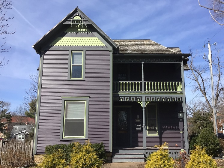 2-story wooden home painted dark purple.  Has a front porch with another balcony/porch above it.  Example of Folk Victorian style.