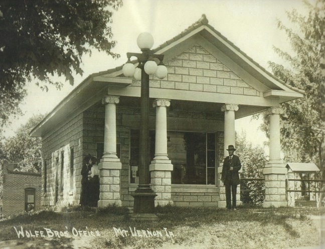 1920 black and white photo of Dr. Wolfe's office, a small 1 story stone building with a large overhanging open porch over a huge open window.  There is a man standing on the right and one or two women on the far left.
