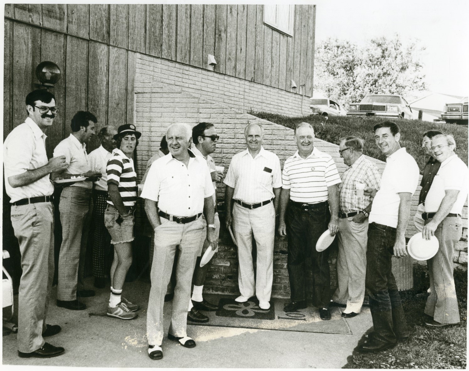 Black and white photo of a group of men holding paper plates standing behind a barn.