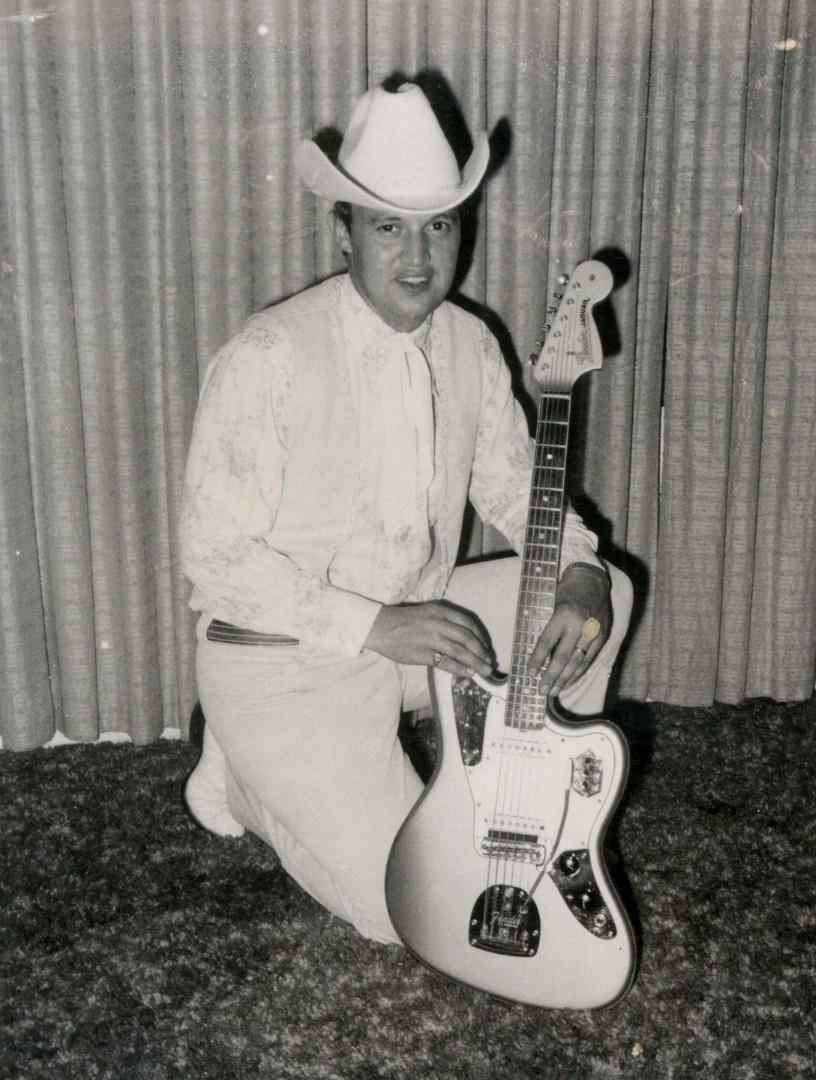 Photo of John Krob wearing a white cowboy outfit kneeling beside his guitar.